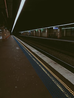 A dimly lit train station platform with yellow and blue tactile paving strips. There are multiple tracks visible, and signage indicating 'Mind The Gap' near the edge of the platform. Overhead lighting casts a linear illumination over the area, and there are empty benches along the opposite platform.