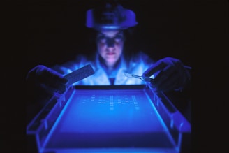A person wearing protective gloves and a face shield is examining a glowing blue surface, possibly a gel electrophoresis apparatus, in a dimly lit lab environment. The scene is illuminated by the blue light, highlighting the focused expression and tools held in the hands.