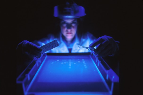 A person wearing protective gloves and a face shield is examining a glowing blue surface, possibly a gel electrophoresis apparatus, in a dimly lit lab environment. The scene is illuminated by the blue light, highlighting the focused expression and tools held in the hands.