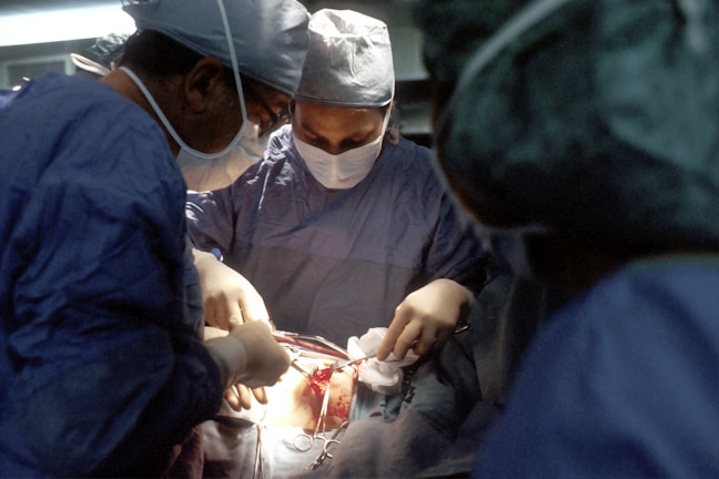 A group of surgeons are performing an operation in a well-equipped, sterile environment. The focus is on the patient's open chest cavity where the surgical procedure is taking place. Surgeons are wearing blue surgical scrubs, masks, and caps. Bright lighting is used to illuminate the area being operated on.
