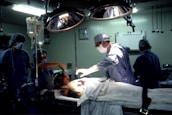 Medical professionals are performing surgery in an operating room equipped with overhead lights and medical equipment. IV bags are visible, and the team is focused on the patient lying on the operating table.