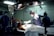 Medical professionals are performing surgery in an operating room equipped with overhead lights and medical equipment. IV bags are visible, and the team is focused on the patient lying on the operating table.