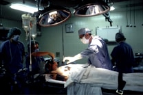 Medical professionals are performing surgery in an operating room equipped with overhead lights and medical equipment. IV bags are visible, and the team is focused on the patient lying on the operating table.