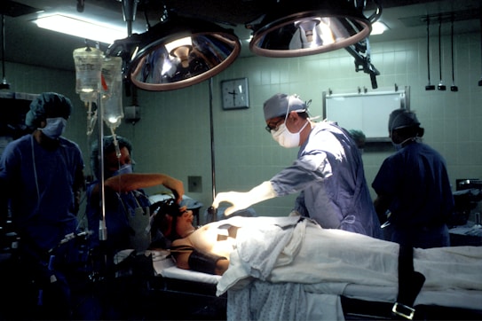 Medical professionals are performing surgery in an operating room equipped with overhead lights and medical equipment. IV bags are visible, and the team is focused on the patient lying on the operating table.