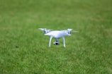 A white quadcopter drone is hovering closely above a lush, green grassy field.