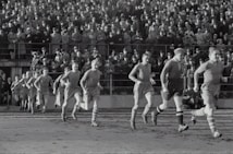 A group of football players in vintage uniforms is seen running on a grass field, with a large crowd of spectators in the background. The crowd is dressed in old-fashioned attire, indicating that this is a historical sports event.