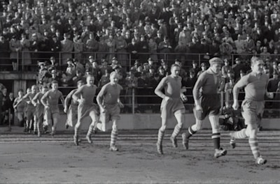 A group of football players in vintage uniforms is seen running on a grass field, with a large crowd of spectators in the background. The crowd is dressed in old-fashioned attire, indicating that this is a historical sports event.