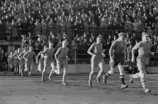 A group of football players in vintage uniforms is seen running on a grass field, with a large crowd of spectators in the background. The crowd is dressed in old-fashioned attire, indicating that this is a historical sports event.