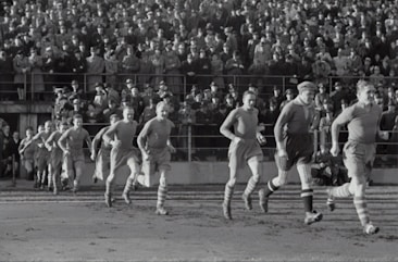 A group of football players in vintage uniforms is seen running on a grass field, with a large crowd of spectators in the background. The crowd is dressed in old-fashioned attire, indicating that this is a historical sports event.