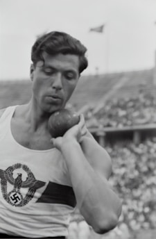 A black and white photo of a male athlete preparing to throw a shot put. The focus is on the muscular build of the athlete and the concentration on his face. The athlete is wearing a tank top with an emblem printed on it, and a stadium with spectators is visible in the background.