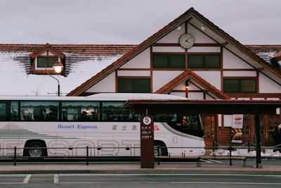 A modern bus labeled 'Resort Express' is parked in front of a building with traditional architecture featuring a triangular roof with red tiles and white walls. A clock is displayed prominently on the building. The roof has some snow, and there is a streetlamp with a vintage design next to the bus.