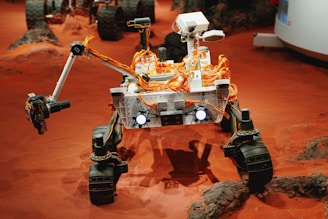 A robotic rover with a white body and numerous orange cables is positioned on a reddish, rocky surface reminiscent of Martian terrain. The rover has large wheels adapted for rough surfaces, and a mechanical arm equipped with instruments. The background features other machinery and a landscape that appears barren and extraterrestrial.