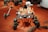A robotic rover with a white body and numerous orange cables is positioned on a reddish, rocky surface reminiscent of Martian terrain. The rover has large wheels adapted for rough surfaces, and a mechanical arm equipped with instruments. The background features other machinery and a landscape that appears barren and extraterrestrial.