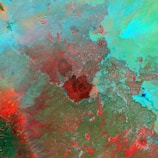 A satellite image showcasing diverse landforms with varying colors. Red and brown regions appear to highlight elevation or different terrain, while greens and blues may represent vegetation or bodies of water. The image has a mix of organic patterns indicating natural landscapes.