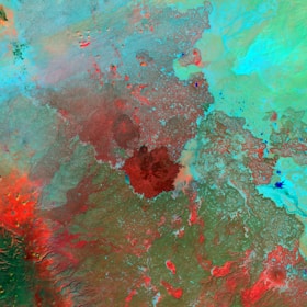 A satellite image showcasing diverse landforms with varying colors. Red and brown regions appear to highlight elevation or different terrain, while greens and blues may represent vegetation or bodies of water. The image has a mix of organic patterns indicating natural landscapes.