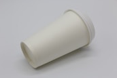 A plain white, disposable paper cup with a lid is lying on its side against a plain background.