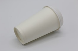 A plain white, disposable paper cup with a lid is lying on its side against a plain background.