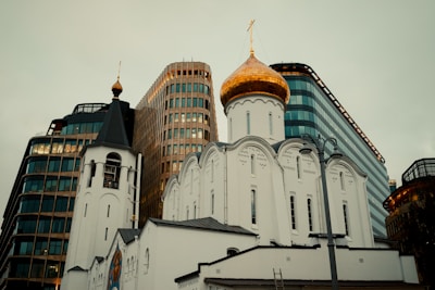 A white Orthodox church with a gold dome stands prominently in front of modern office buildings. The church features ornate architectural details and a tall bell tower. The backdrop of contemporary glass and steel structures creates a striking contrast between traditional and modern elements.