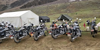 A row of multiple motorcycles parked on a grassy terrain near large camping tents. The setting appears to be outdoors, possibly in a mountainous region, with rocky slopes in the background.