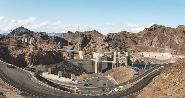 A group of engineers examining blueprints beside a large earth dam under construction on a sunny day.