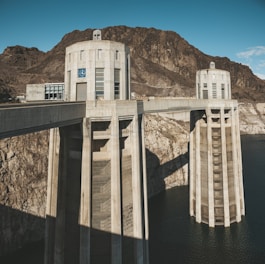 The image features a massive concrete dam structure with two circular towers, both with clock faces. The dam is situated in a rocky desert landscape with a body of water visible below. The sky is clear and blue, contributing to the stark, dramatic appearance of the structure against the rugged terrain.
