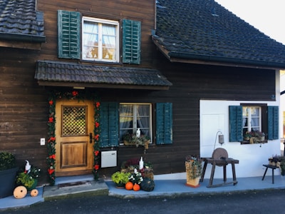 A rustic house with a wooden facade features a wooden door decorated with greenery and red ornaments. The windows have green shutters and are adorned with lace curtains. In front of the house, there are potted plants, pumpkins, and flowers arranged neatly. A small, antique wooden table with a hanging lantern is placed near the entrance.