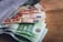 Several euro banknotes are partially protruding from an open white envelope placed on a wooden surface. The denominations of the visible banknotes include 5, 10, and 20 euros.