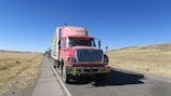 A red cargo truck with a sign for a gas company is parked on a rural road with a clear blue sky in the background. The truck is followed by several other vehicles in a queue. The landscape around the road is dry with patches of grass and distant hills.