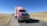 A red cargo truck with a sign for a gas company is parked on a rural road with a clear blue sky in the background. The truck is followed by several other vehicles in a queue. The landscape around the road is dry with patches of grass and distant hills.