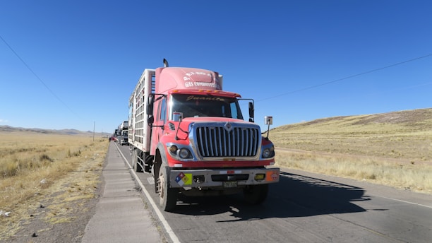 A red cargo truck with a sign for a gas company is parked on a rural road with a clear blue sky in the background. The truck is followed by several other vehicles in a queue. The landscape around the road is dry with patches of grass and distant hills.
