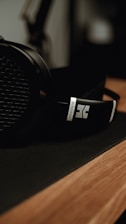 A close-up of a pair of over-ear headphones resting on a smooth wooden surface. The focus is on the branding on the headband, with a soft, dark, and moody background.