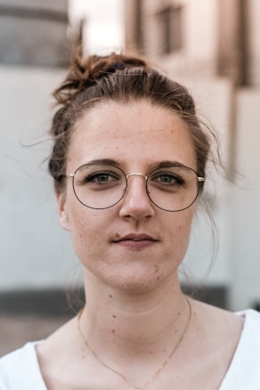 A person with tied-back hair and round glasses is looking directly at the camera. The background is blurred, emphasizing the subject's face. The lighting is even, highlighting facial features.