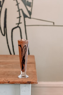 A tall glass filled with a creamy chocolate drink sits on a rustic wooden table. The glass features artistic swirls of dark chocolate sauce visible through the sides. In the background, there is abstract black and white wall art.