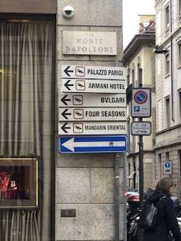 Street signs on a stone wall direct people to various high-end hotels and brands, including Palazzo Parigi, Armani Hotel, Bulgari, Four Seasons, and Mandarin Oriental. A woman's silhouette is seen walking near the signs. Behind the signs, a building with a decorative glass element and a jewelry display is partially visible.