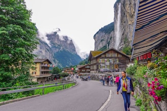 A picturesque village scene set against a stunning backdrop of towering cliffs and a cascading waterfall. Traditional wooden houses line the street, with colorful flags and flowers adding charm to the scene. People are walking along the road, some taking pictures, as the clouds hover over the distant mountains.