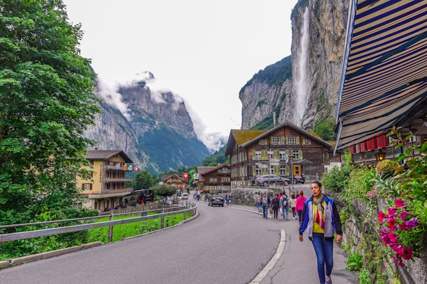 A picturesque village scene set against a stunning backdrop of towering cliffs and a cascading waterfall. Traditional wooden houses line the street, with colorful flags and flowers adding charm to the scene. People are walking along the road, some taking pictures, as the clouds hover over the distant mountains.