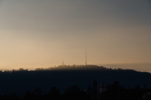 A silhouette of a hilly landscape with a prominent radio tower standing out against a muted, golden sky. The horizon is softly lit, with trees and buildings forming a dark foreground.
