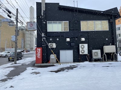 A black building with air conditioning units mounted on the side is visible, surrounded by snow. The street is covered in a thin layer of snow, and utility poles with power lines run alongside the road. A Coca-Cola vending machine is positioned near the building. On the left, several vehicles drive on the snowy street while light snow appears to be falling.