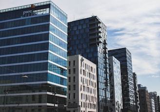 A series of modern high-rise office buildings with reflective glass facades stand against a partly cloudy sky. The architecture features a mix of rectangular shapes and varied designs.