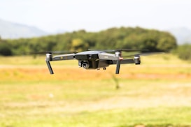 A drone with black and gray colors hovers in mid-air against a blurred natural background. The drone features four propellers and a central camera mounted on its body.