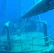 A submerged shipwreck with rust-covered metal surfaces is visible underwater. Barnacles and marine growths clinging to the rails and structures create a sense of abandonment. The deep blue water surrounds the vessel, giving a serene yet eerie atmosphere.