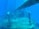 A submerged shipwreck with rust-covered metal surfaces is visible underwater. Barnacles and marine growths clinging to the rails and structures create a sense of abandonment. The deep blue water surrounds the vessel, giving a serene yet eerie atmosphere.