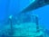A submerged shipwreck with rust-covered metal surfaces is visible underwater. Barnacles and marine growths clinging to the rails and structures create a sense of abandonment. The deep blue water surrounds the vessel, giving a serene yet eerie atmosphere.