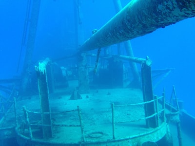 A submerged shipwreck with rust-covered metal surfaces is visible underwater. Barnacles and marine growths clinging to the rails and structures create a sense of abandonment. The deep blue water surrounds the vessel, giving a serene yet eerie atmosphere.