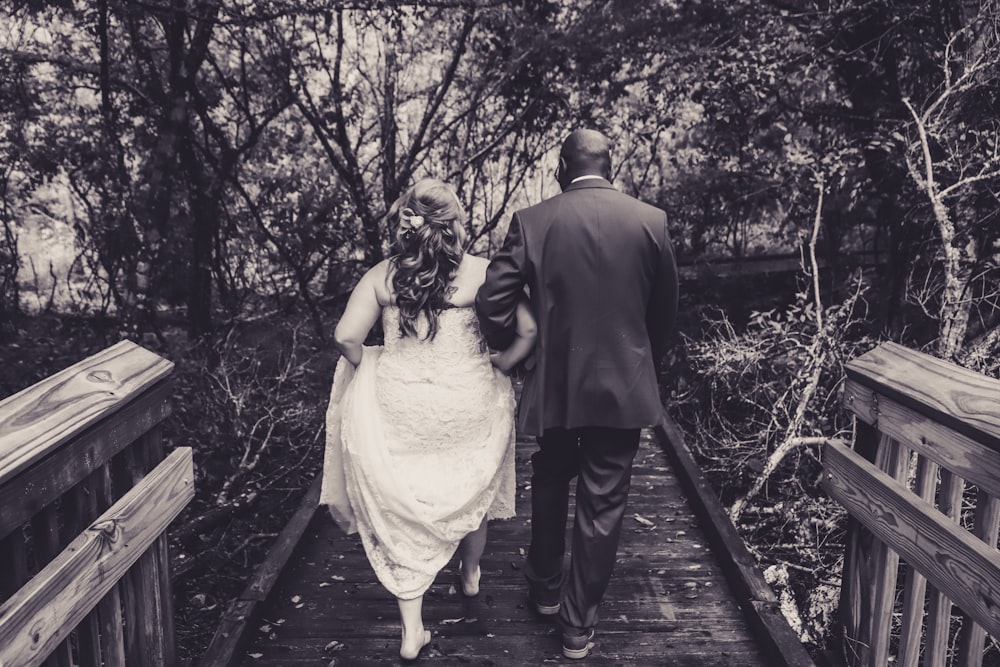 grayscale photography of woman in dress and man in tuxedo walking on wooden bridge