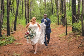 A couple walking joyfully through a wooded area, with the woman in a white wedding dress holding a bouquet of red and white flowers and greenery. The man, wearing a dark suit, smiles as he holds the train of her dress. Tall trees surround them, and the forest floor is covered with pine needles.
