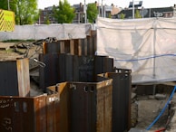 A construction site with metal sheet piles arranged in a circle in the foreground. The background shows a fenced-off area with a white tarp and a 'Hitachi' branded sign. Buildings and trees are visible in the distance.