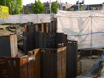 A construction site with metal sheet piles arranged in a circle in the foreground. The background shows a fenced-off area with a white tarp and a 'Hitachi' branded sign. Buildings and trees are visible in the distance.
