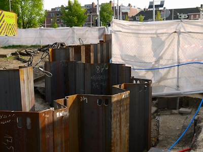 A construction site with metal sheet piles arranged in a circle in the foreground. The background shows a fenced-off area with a white tarp and a 'Hitachi' branded sign. Buildings and trees are visible in the distance.