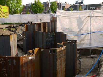 A construction site with metal sheet piles arranged in a circle in the foreground. The background shows a fenced-off area with a white tarp and a 'Hitachi' branded sign. Buildings and trees are visible in the distance.